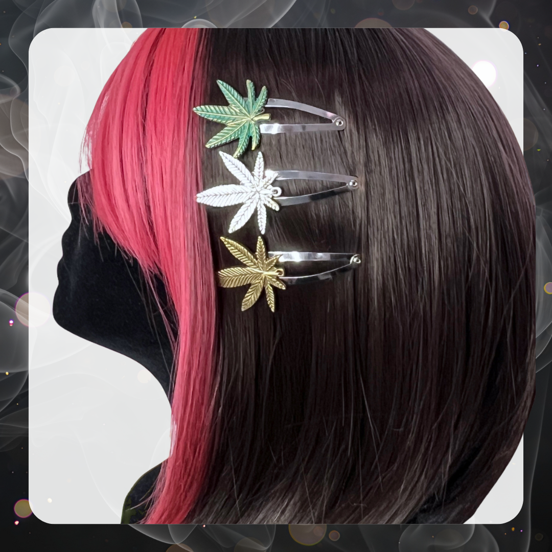 Picture of a mannequin head with brown and red hair wearing three different marijuana hair clips on the side of their hair. The three hair clips are first a green weed leaf, second is a silver leaf, and third is a bronze leaf. All are silver snap hair clips.