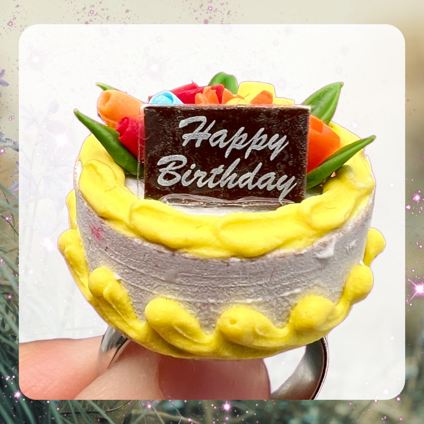 A picture of a tiny miniature birthday cake with yellow decorative frost using and flowers in on top. There is a tiny sign on the cake that says Happy Birthday.