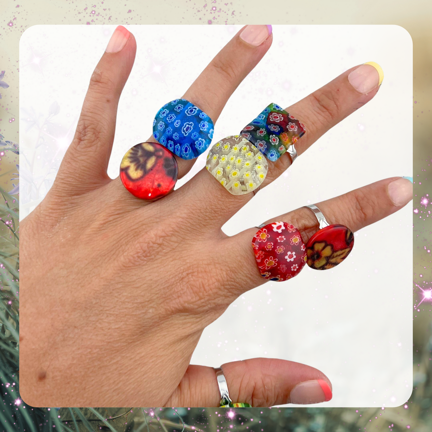 Picture shows a woman’s hand wearing seven variations of rings. The rings are made of glass beads that have a design of tiny flowers inside of them. There is a blue, red, yellow, and multicolor ring.