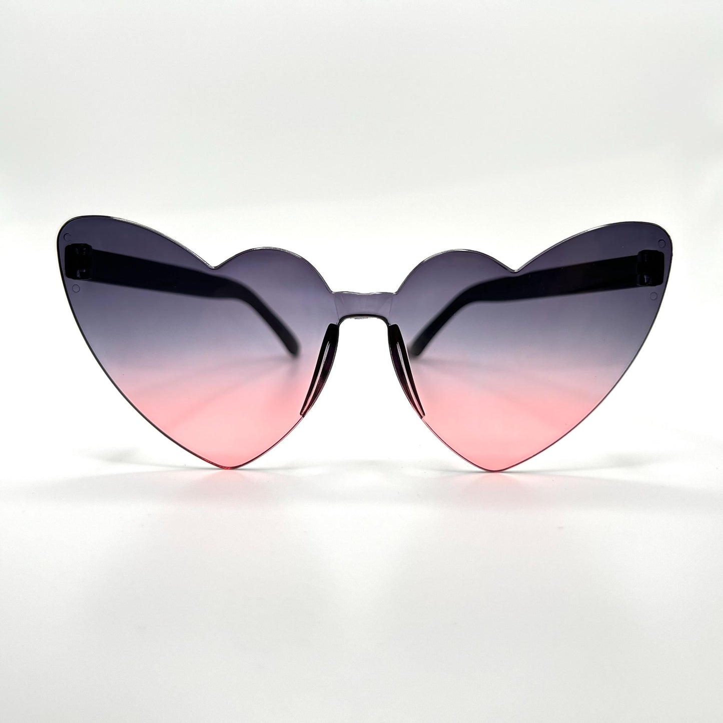 Colorful oversized heart-shaped sunglasses, perfect for festivals and dancing. Lightweight and trendy eyewear for a vibrant and stylish look. Displayed on a table. Front view. Black pink Ombre Cat Eye Heart.