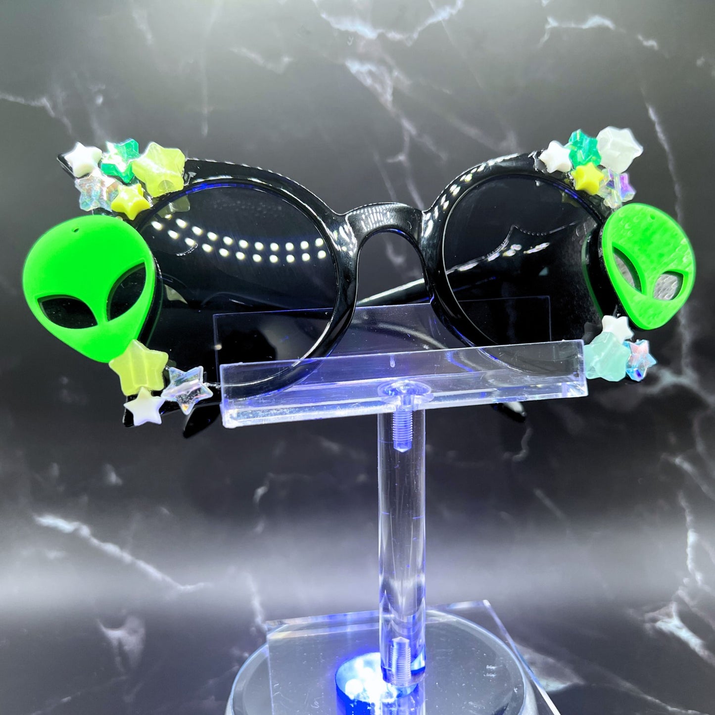 Glow-in-the-dark star festival sunglasses for an out-of-this-world look. Displayed on a clear stand. Front view.