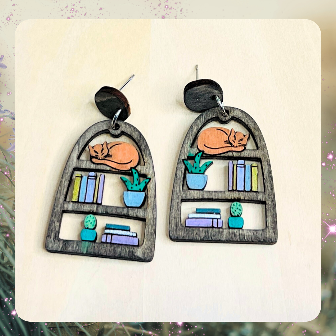 Earrings with an orange kitty cat sleeping on a mini bookshelf with plants. Handmade and hand-painted wood. 