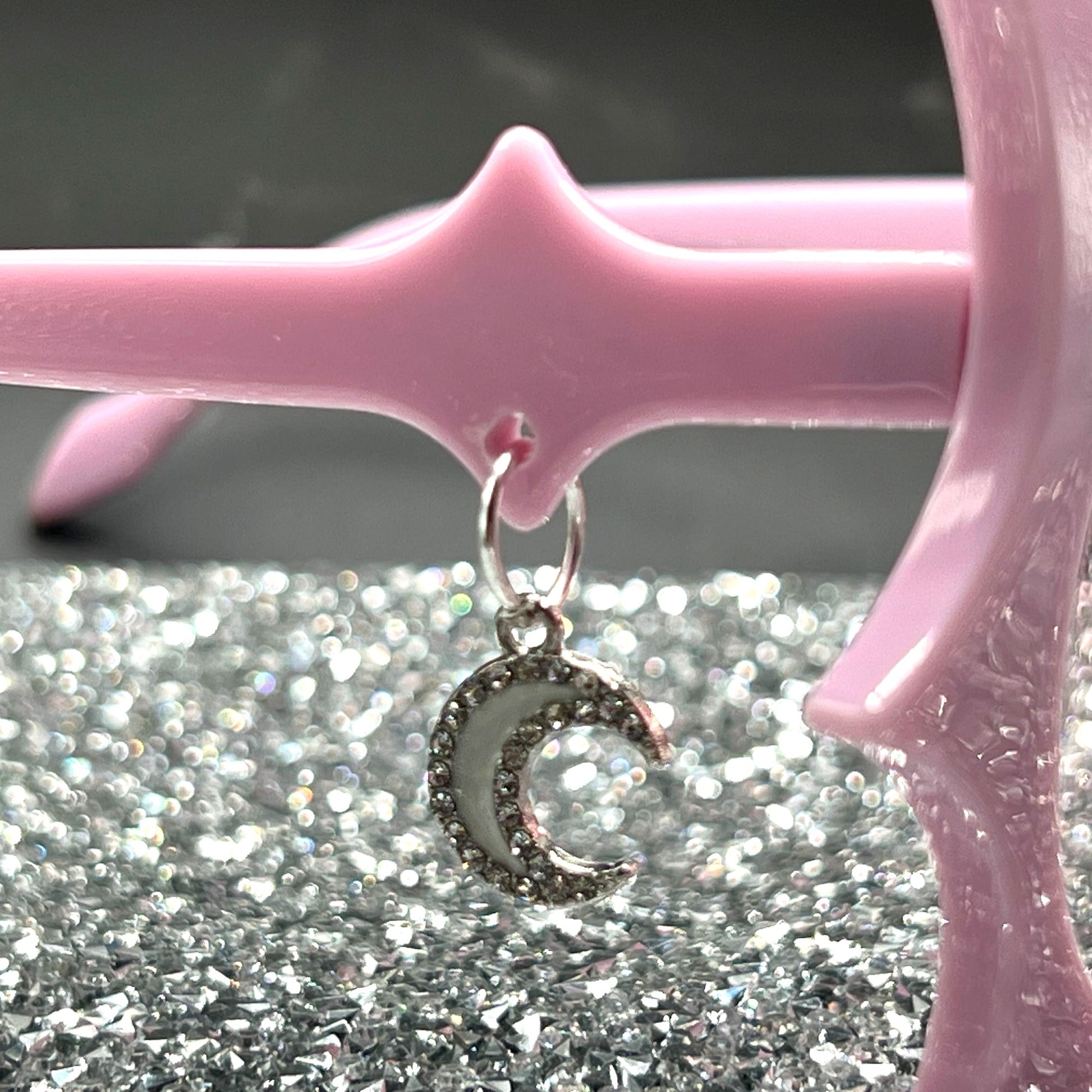 "Pastel pink bat sunglasses with moon charms. Displayed on a glittery table. Close up of the moon charm.