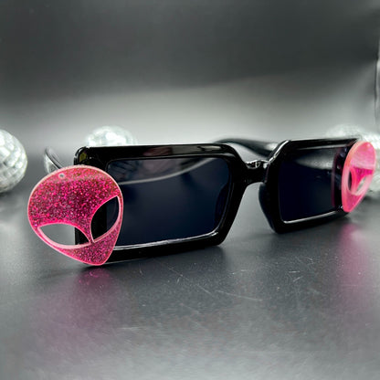 Hot Pink Alien Sunglasses with black frames and glittery pink aliens on sides – a bold and cosmic accessory for festivals and raves. Square/rectangle shape. Displayed on a table. Front view.