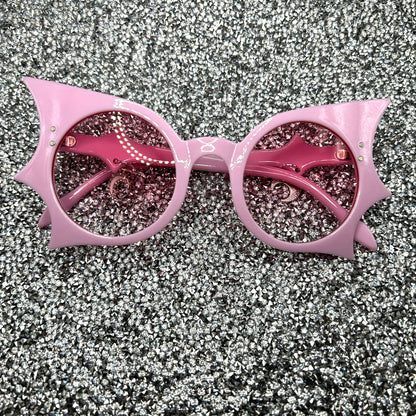 "Pastel pink bat sunglasses with moon charms. Displayed on a glittery table. Laying flat.