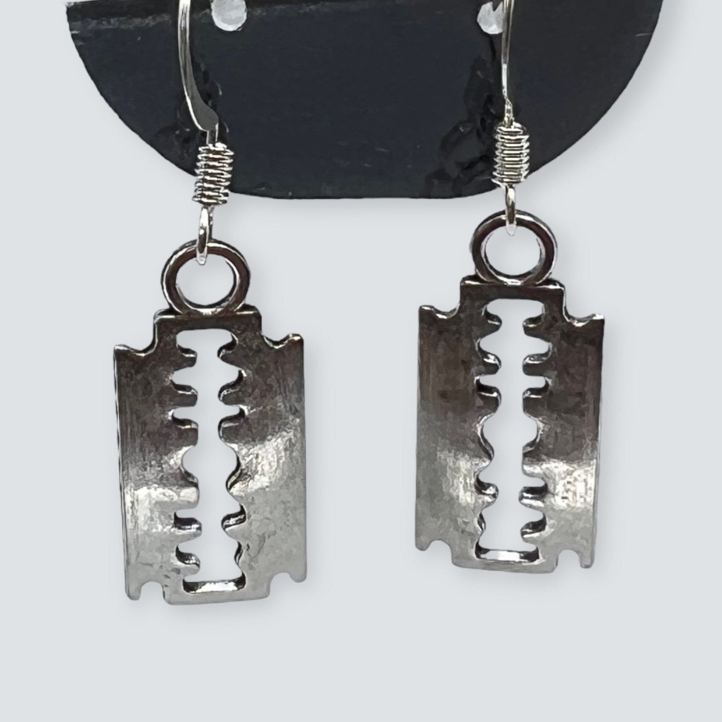 Silver tone razor blade charm earrings, inspired by punk and goth fashion. Displayed on a clear stand.