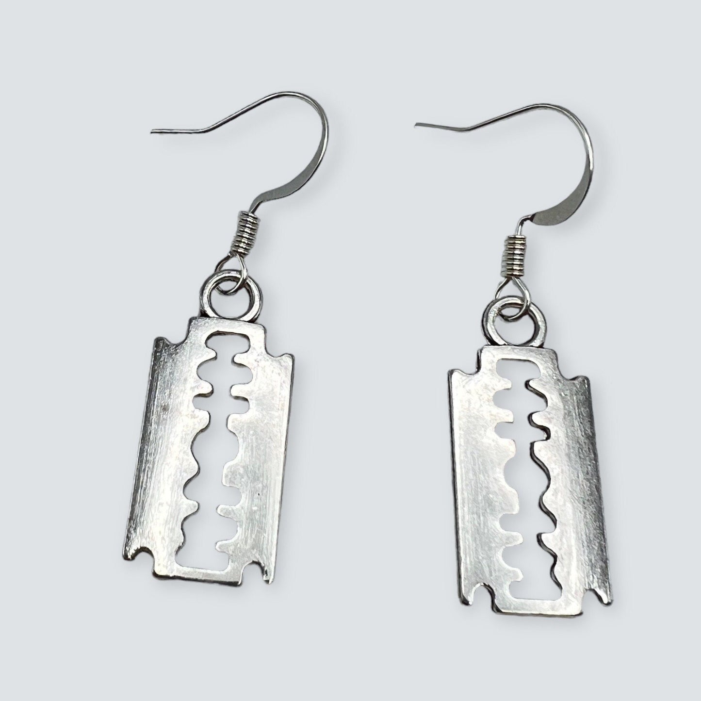 Silver tone razor blade charm earrings, inspired by punk and goth fashion. Displayed on a table.