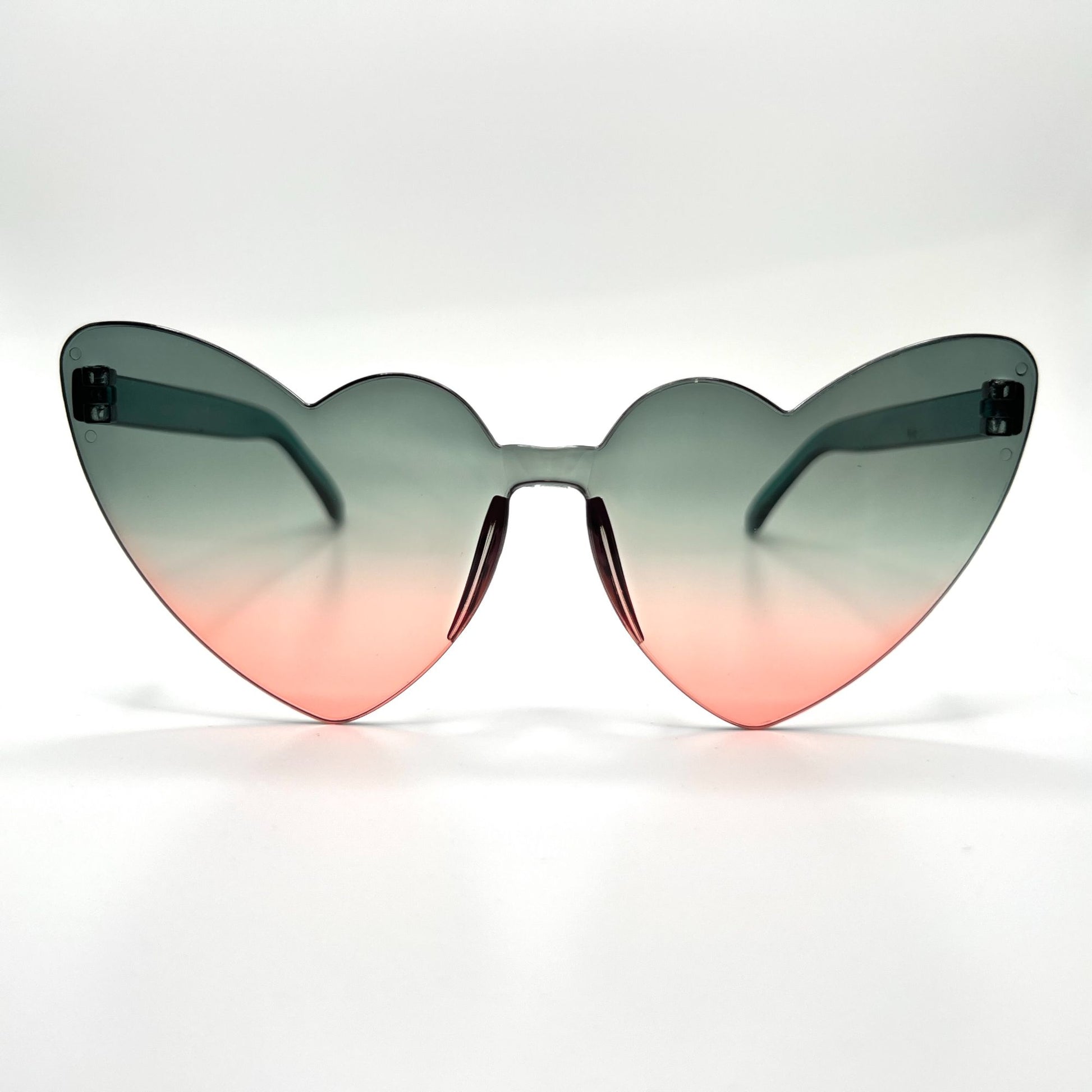 Colorful oversized heart-shaped sunglasses, perfect for festivals and dancing. Lightweight and trendy eyewear for a vibrant and stylish look. Displayed on a table. Front view. Green Pink Ombre Cat Eye Heart.