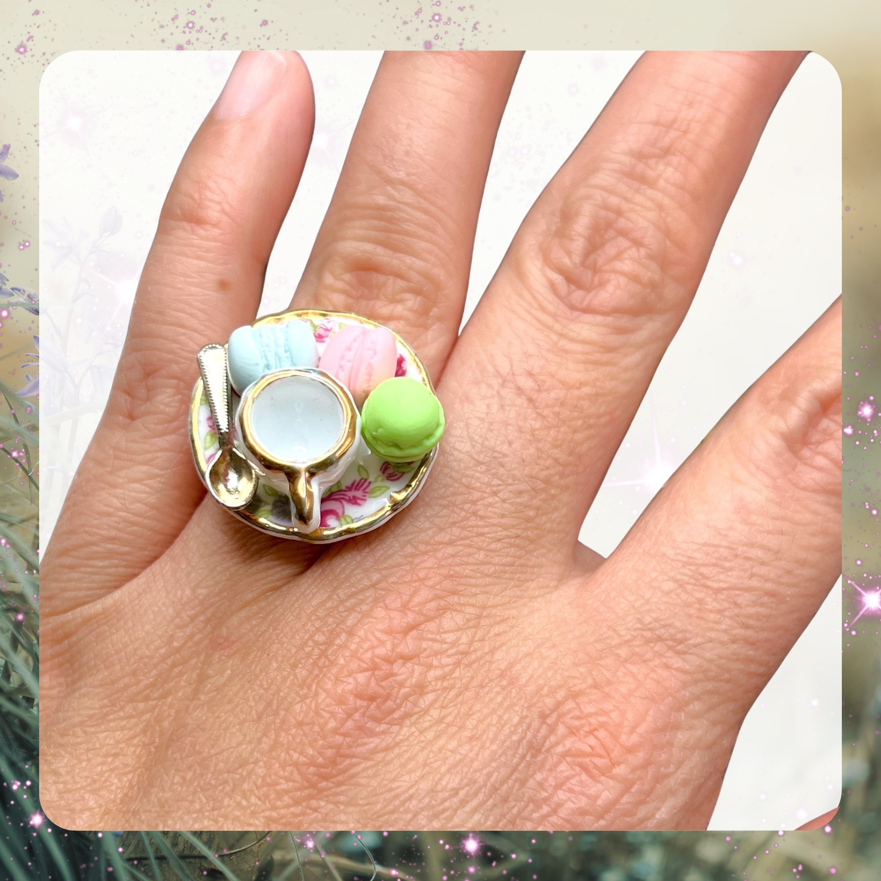 Picture of a woman’s hand wearing a large gaudy statement ring that is a porcelain dollhouse plate with a tiny miniature tea cup and spoon on it plus 3 tiny mini macarons in pastel green, pastel pink, and pastel blue.