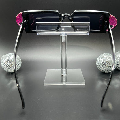 Hot Pink Alien Sunglasses with black frames and glittery pink aliens on sides – a bold and cosmic accessory for festivals and raves. Square/rectangle shape. Displayed on a clear stand. Back view.