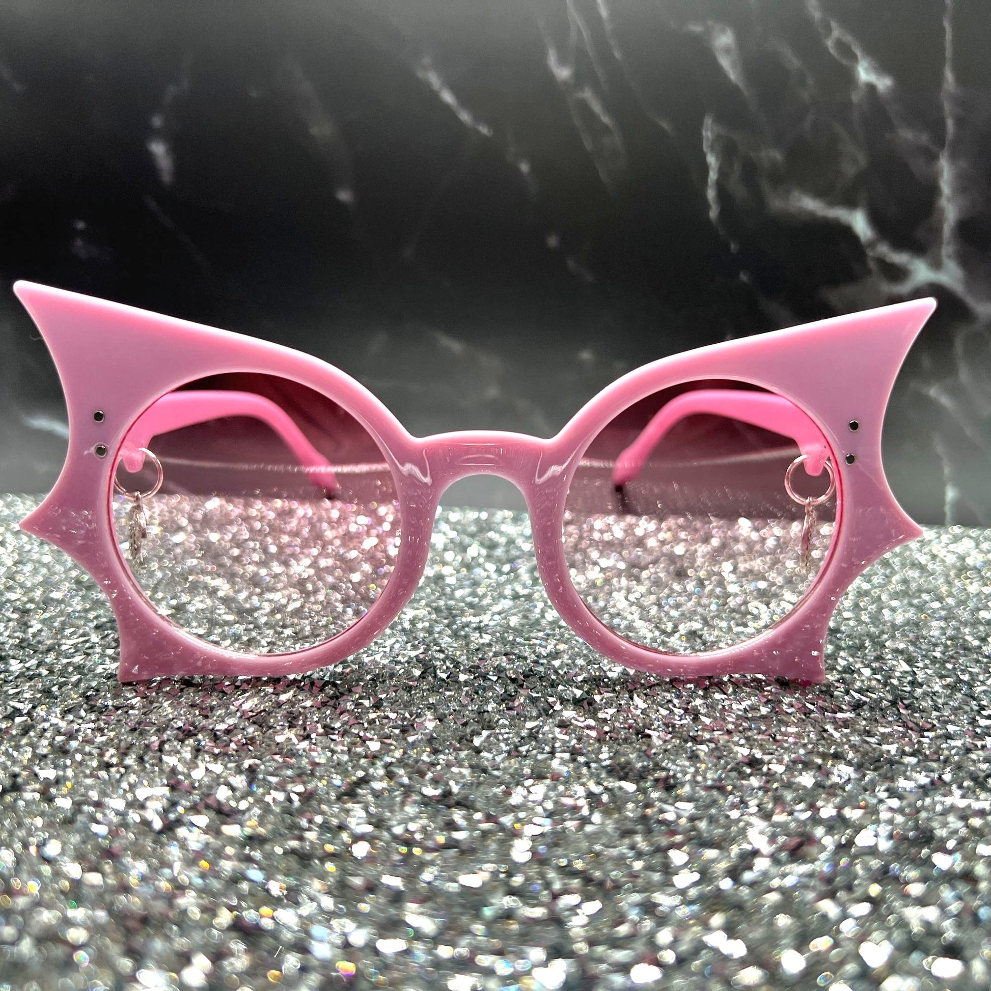 "Pastel pink bat sunglasses with moon charms. Displayed on a glittery table.