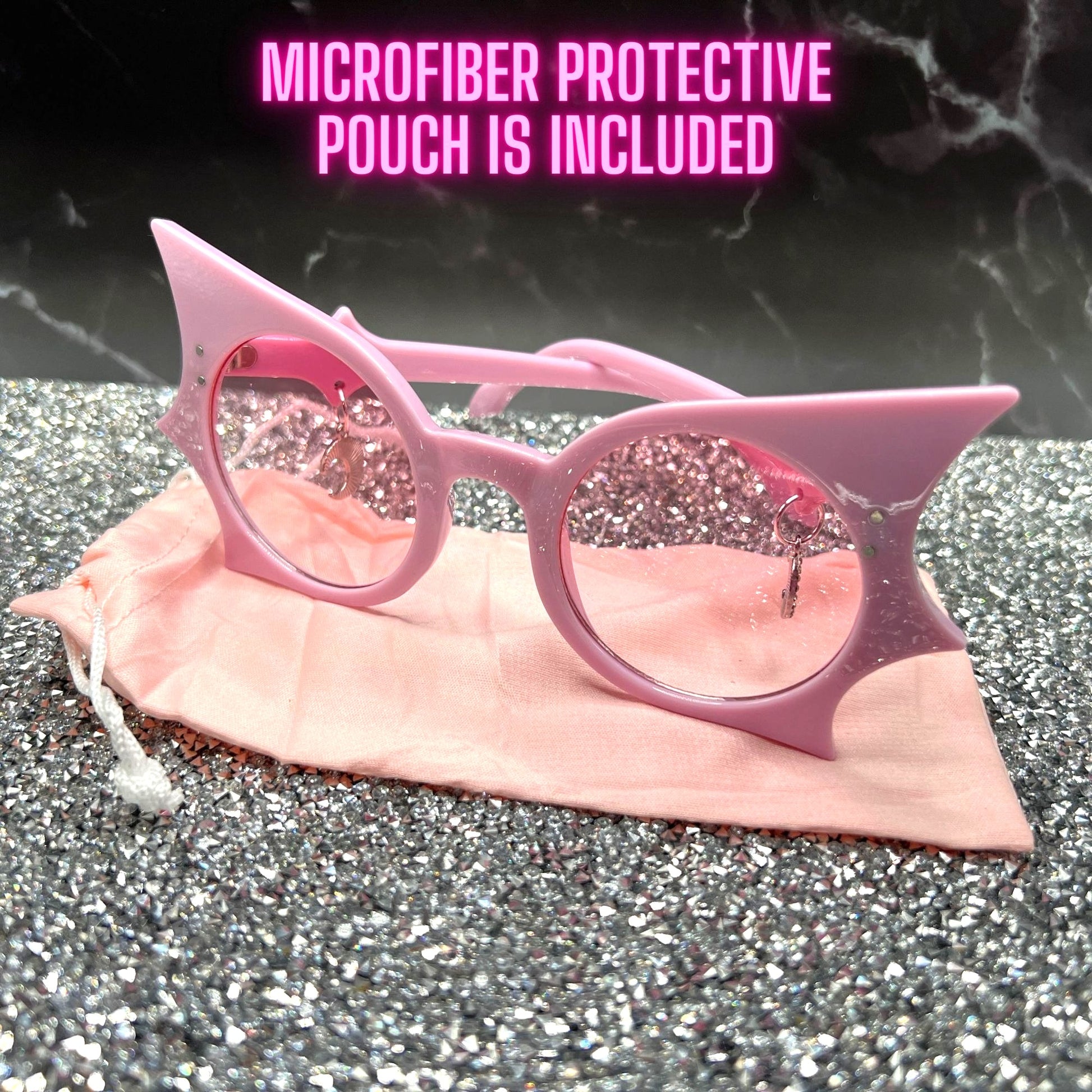 "Pastel pink bat sunglasses with moon charms. Displayed on a glittery table with a pink microfiber cloth pouch that is included.