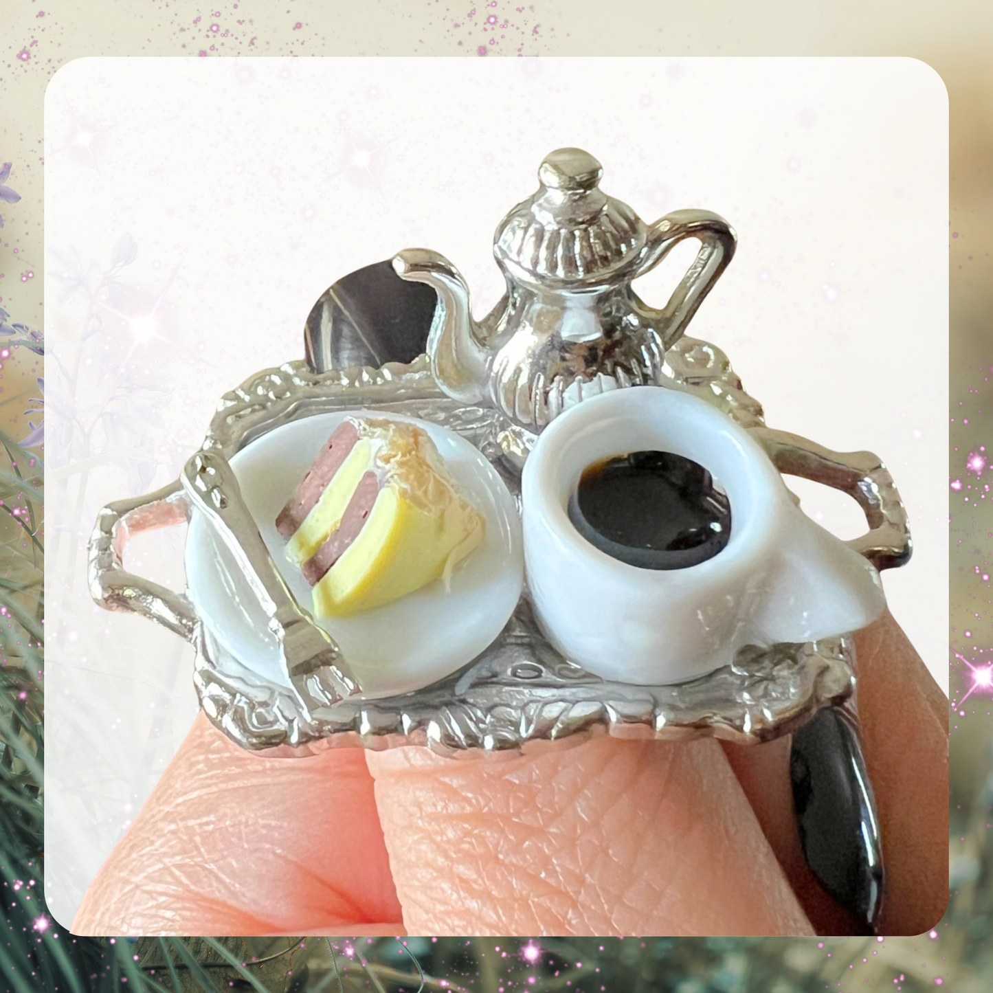 Picture shows a miniature silver metal dollhouse tea tray set with a mini silver tea kettle, a white mug with brown tea inside, and tiny white plate with a slice of yellow and brown cake on it with a tiny fork, all made into a cocktail ring. 