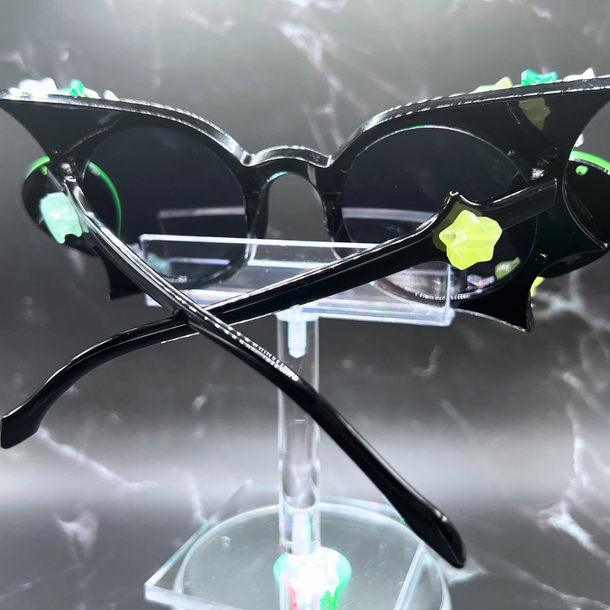 Glow-in-the-dark star festival sunglasses for an out-of-this-world look. Displayed on a clear stand. Side view.