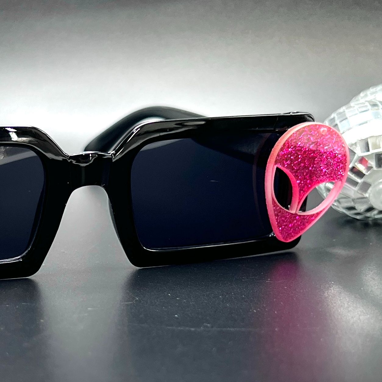 Hot Pink Alien Sunglasses with black frames and glittery pink aliens on sides – a bold and cosmic accessory for festivals and raves. Square/rectangle shape. Displayed on a table. Front view.