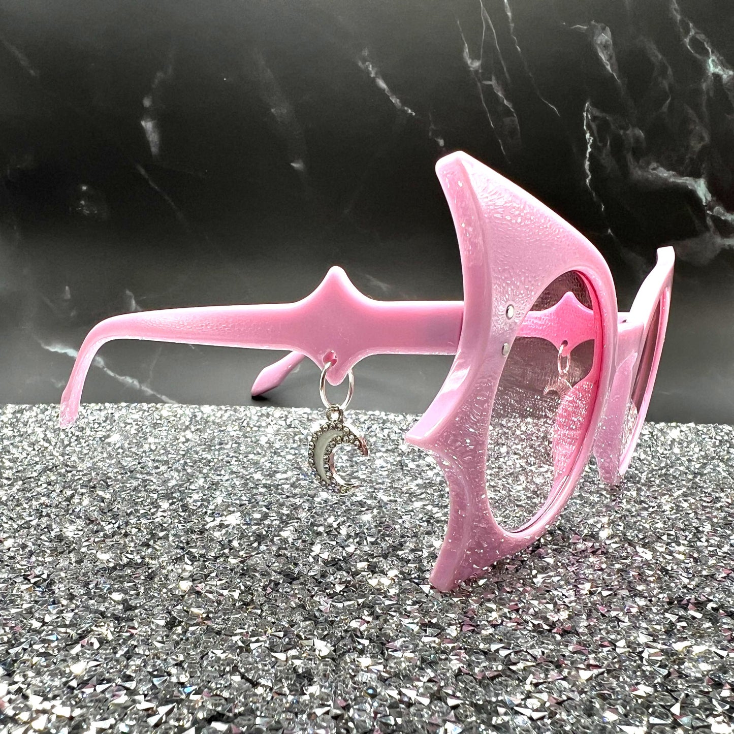 "Pastel pink bat sunglasses with moon charms. Displayed on a glittery table. Side view.