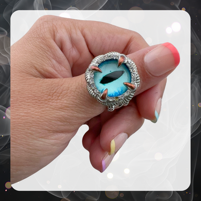 Picture is a woman’s hand showing the thumb with a large blue dragon eye on a silver tone ring. 