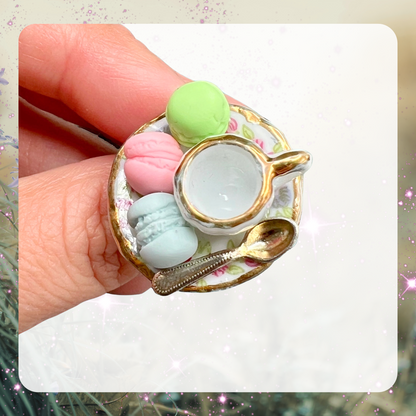 Picture of a woman’s hand wearing a large gaudy statement ring that is a porcelain dollhouse plate with a tiny miniature tea cup and spoon on it plus 3 tiny mini macarons in pastel green, pastel pink, and pastel blue.