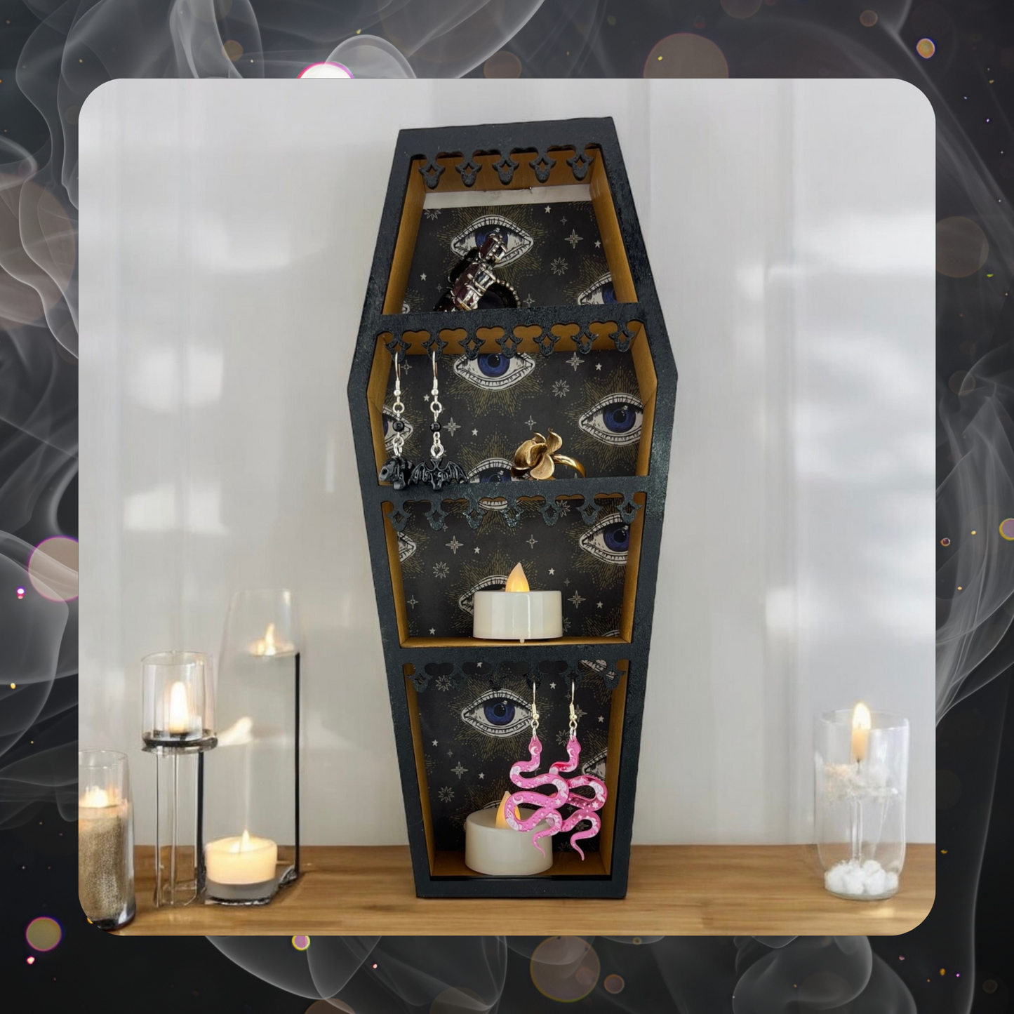 The picture shows a miniature coffin curio shelf on a table that is decorating with candles. The curio shelf has some pink snake earrings hanging from one shelf, some bat earrings on another shelf and various rings and jewelry to show how the shelf can be used. 