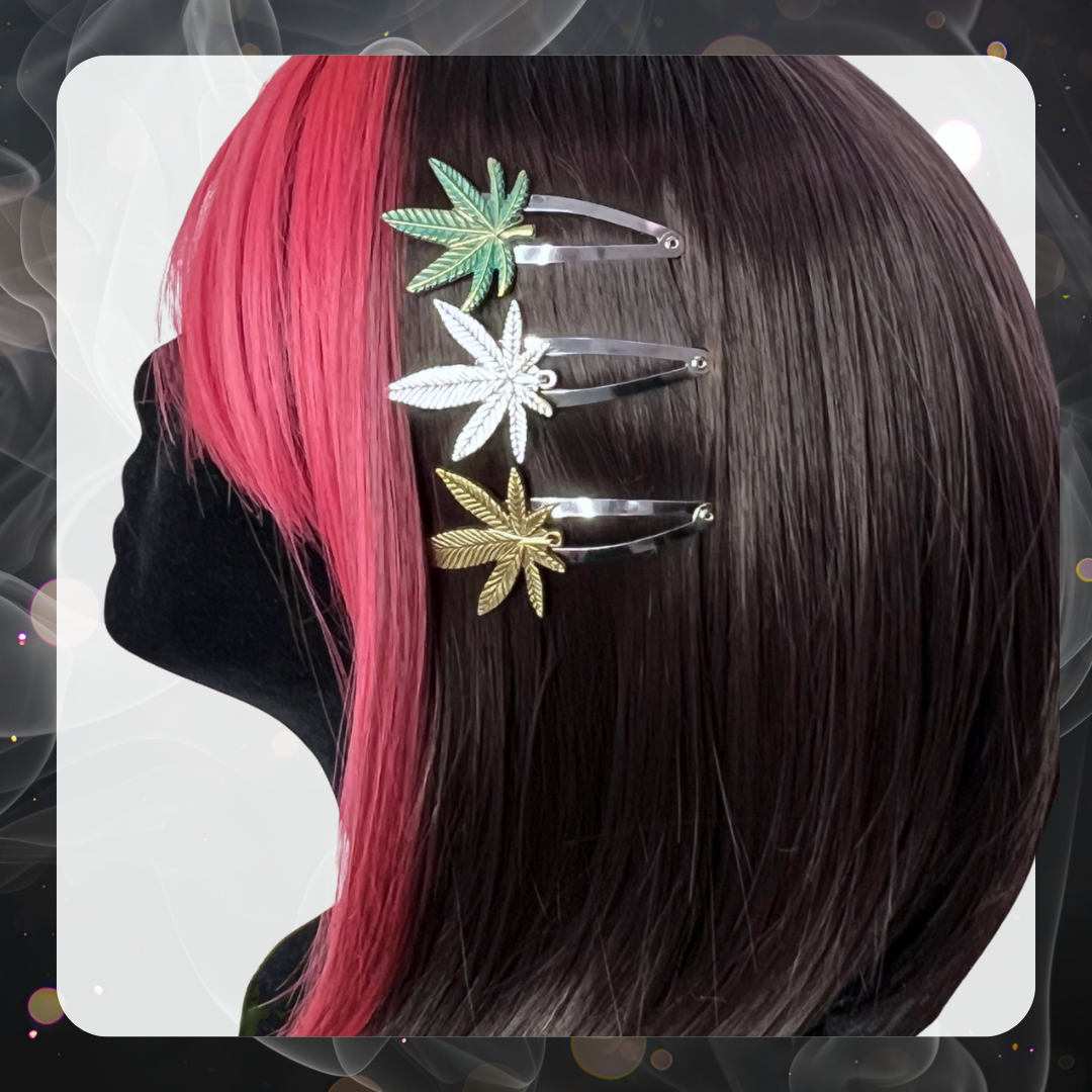 Picture of a mannequin head with brown and red hair wearing three different marijuana hair clips on the side of their hair. The three hair clips are first a green weed leaf, second is a silver leaf, and third is a bronze leaf. All are silver snap hair clips. 