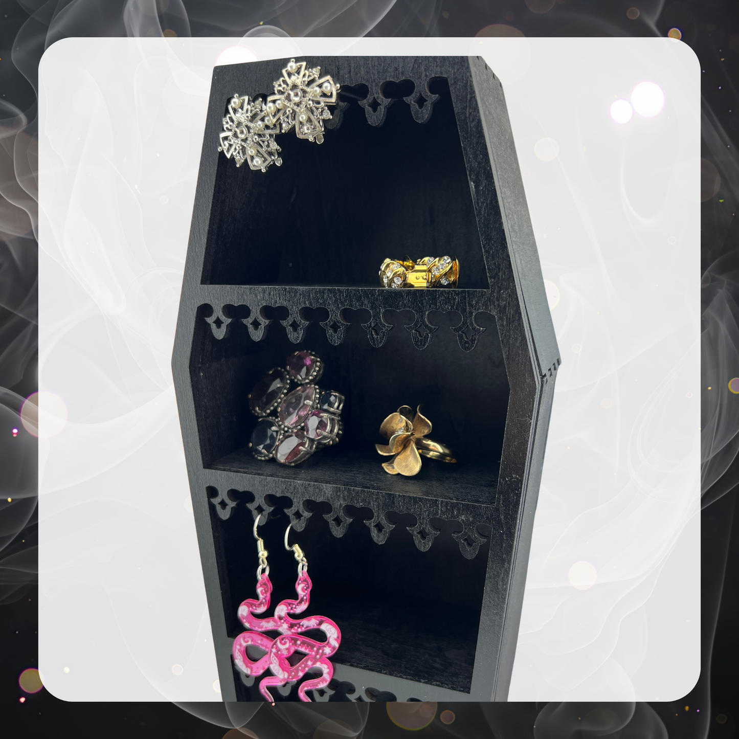 Picture shows a black coffin shaped mini shelf. It is laser cut with ornate edges that have some pink snake earrings hanging on it as well as some rhinestone clip on earrings. The shelves show some costume/cocktail rings.