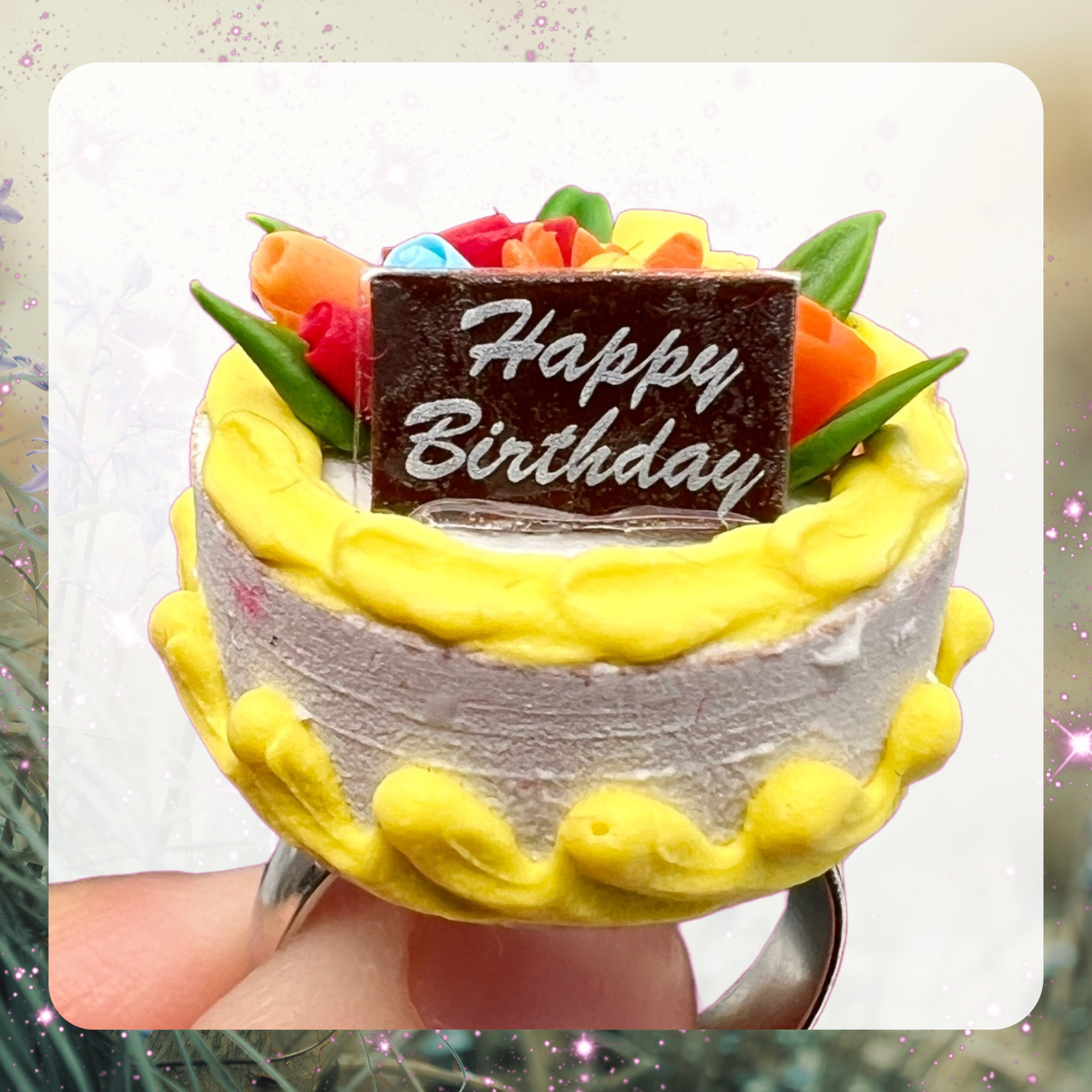 A picture of a tiny miniature birthday cake with yellow decorative frost using and flowers in on top. There is a tiny sign on the cake that says Happy Birthday. 