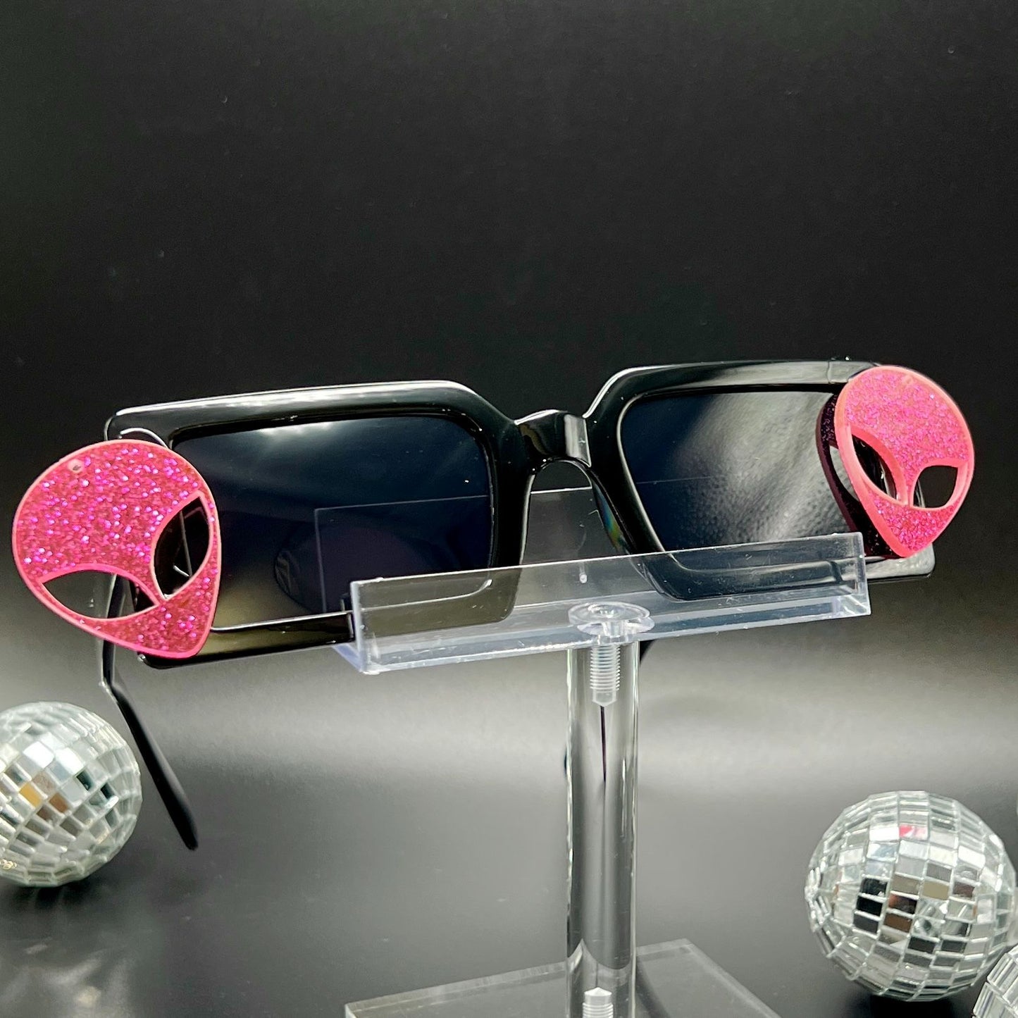 Hot Pink Alien Sunglasses with black frames and glittery pink aliens on sides – a bold and cosmic accessory for festivals and raves. Square/rectangle shape. Displayed on a clear stand. Front view.
