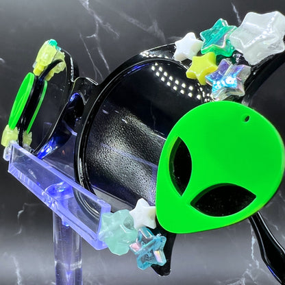 Glow-in-the-dark star festival sunglasses for an out-of-this-world look. Displayed on a clear stand. Side view. Close up of the alien charm.