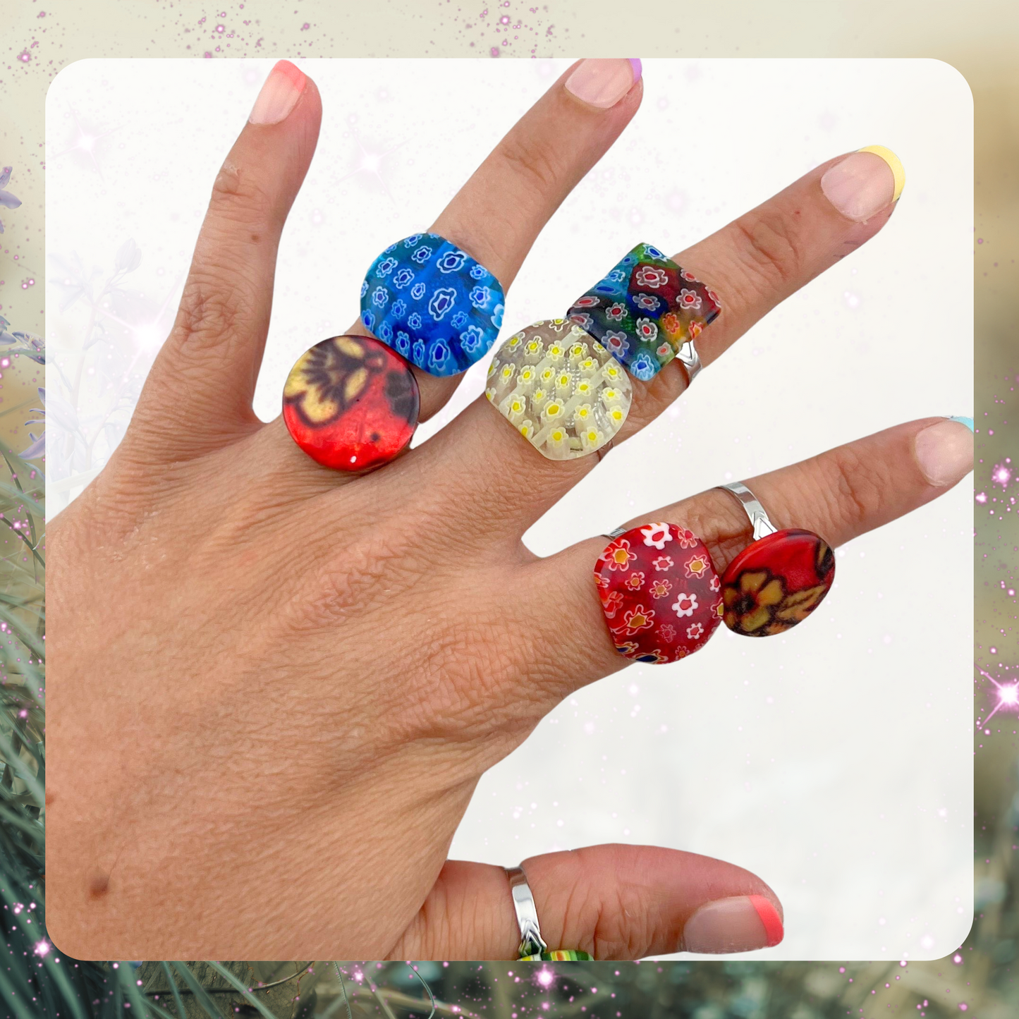 Picture shows a woman’s hand wearing seven variations of rings. The rings are made of glass beads that have a design of tiny flowers inside of them. There is a blue, red, yellow, and multicolor ring. 