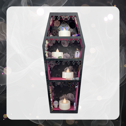 Picture shows a mini coffin shaped curio shelf painted black on the outside and fuchsia pink on the inside as well as with gray tone rose wallpaper on the back. The shelf is adorned with various crystals and tiny trinkets, but there is still room to add you own as well. Each of the four shelves has a battery operated tea light candle on it.  