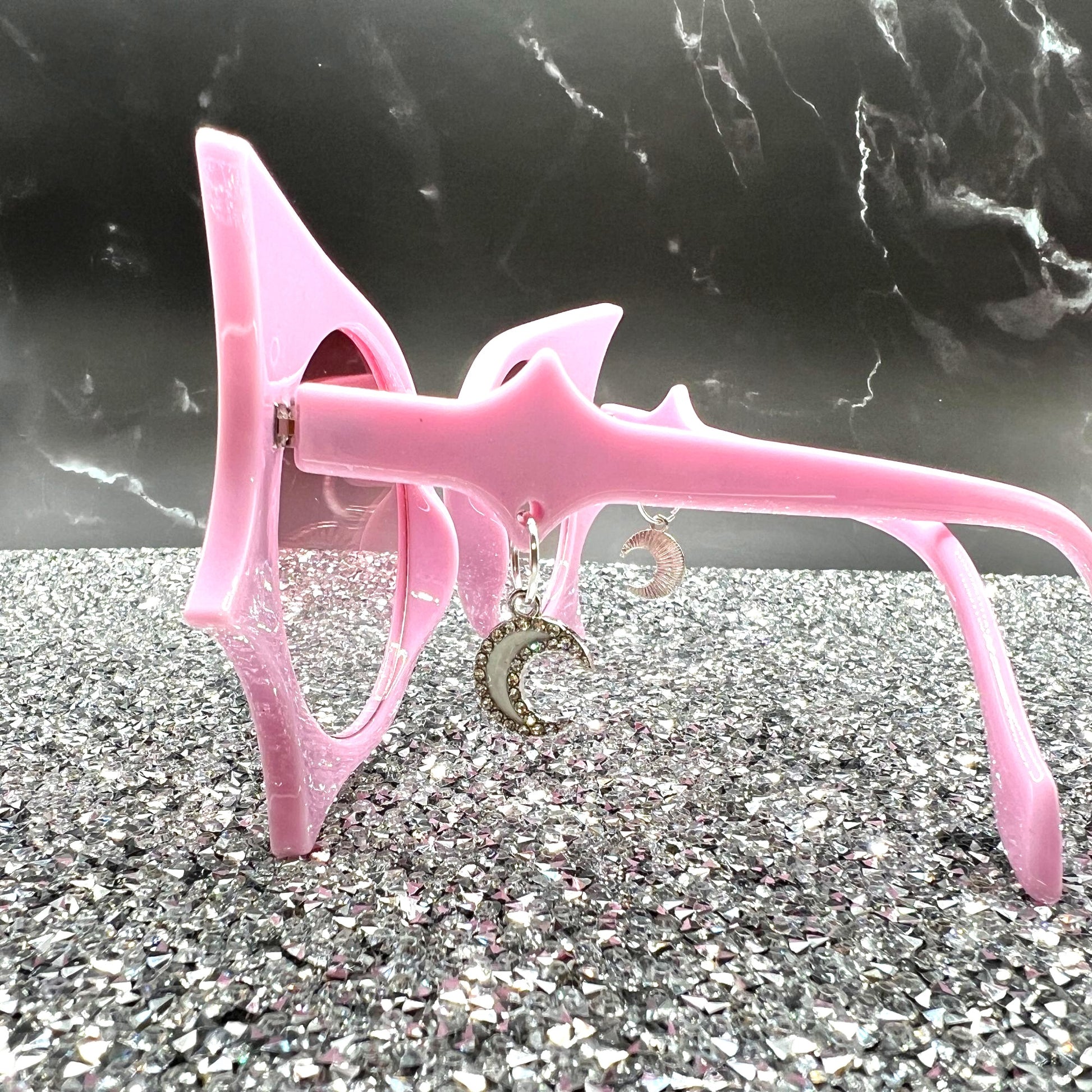 "Pastel pink bat sunglasses with moon charms. Displayed on a glittery table. Side view.