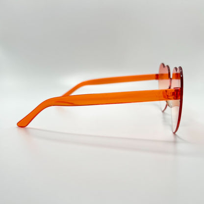 Colorful oversized heart-shaped sunglasses, perfect for festivals and dancing. Lightweight and trendy eyewear for a vibrant and stylish look. Displayed on a table. Side view. Orange Ombre Heart.