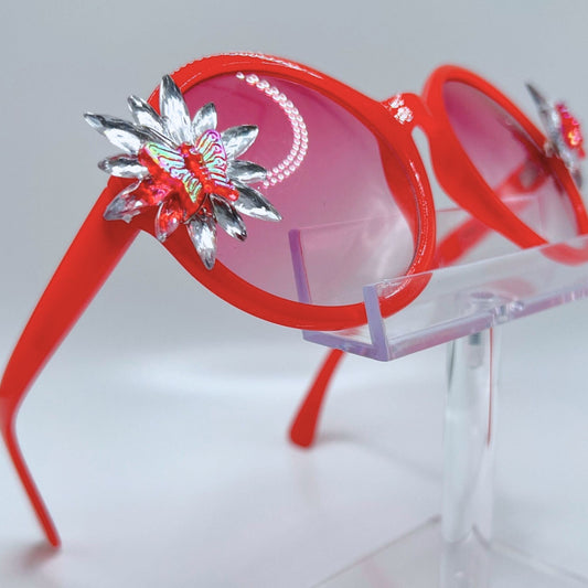 Vibrant red festival sunglasses with rhinestone butterfly bling. Displayed on a clear stand. Front view.