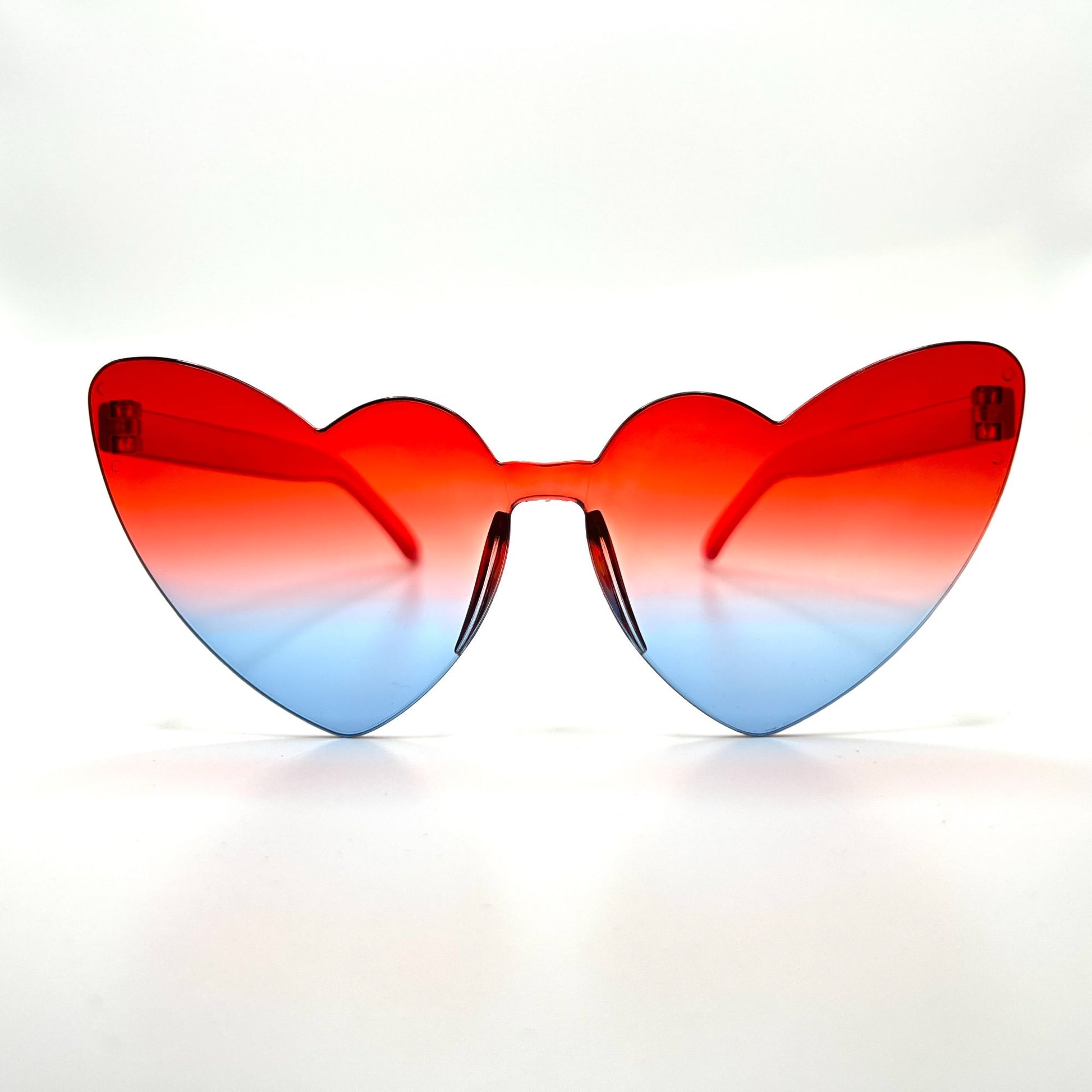 Colorful oversized heart-shaped sunglasses, perfect for festivals and dancing. Lightweight and trendy eyewear for a vibrant and stylish look. Displayed on a table. Front view. Red White Blue Cat Eye Heart.