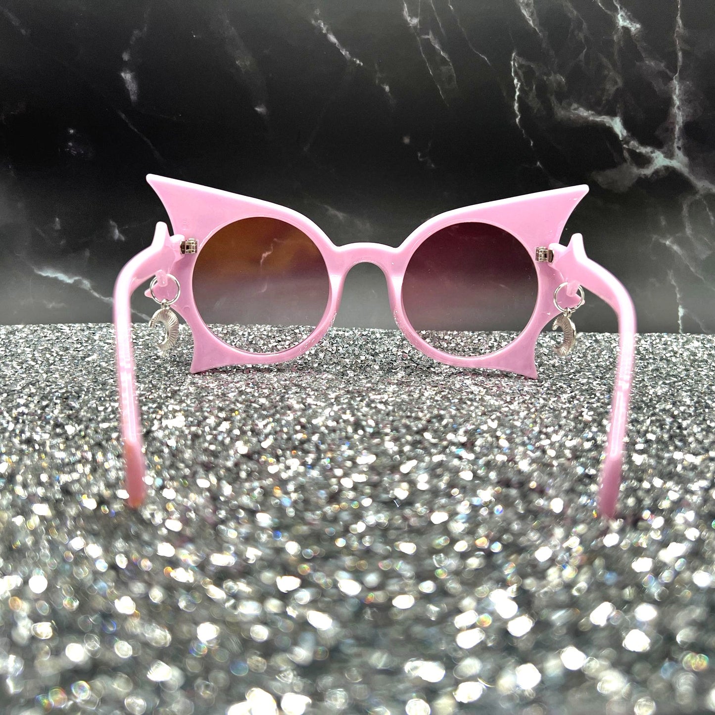 "Pastel pink bat sunglasses with moon charms. Displayed on a glittery table. Back view.
