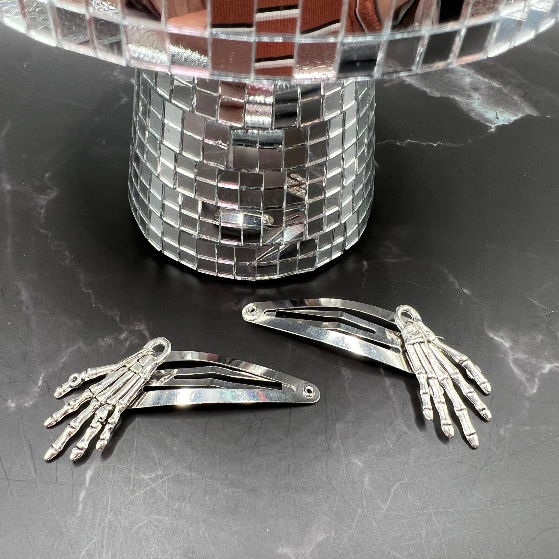 Set of 2 Halloween-themed hair clips with silver metal skeleton hands. Punk rock and gothic girly aesthetics for a bold, cute style. Displayed on a table. Front view.