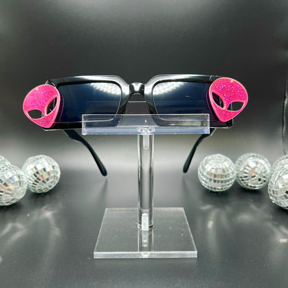 Hot Pink Alien Sunglasses with black frames and glittery pink aliens on sides – a bold and cosmic accessory for festivals and raves. Square/rectangle shape. Displayed on a clear stand. Front view.